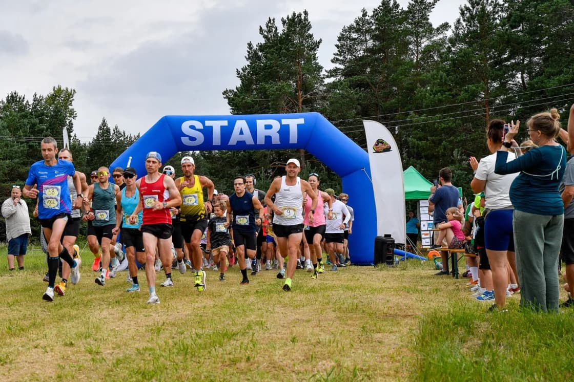 Runners in bright shirts burst out from under a blue inflatable START arch onto grass beneath pine trees, spectators with phones along the side