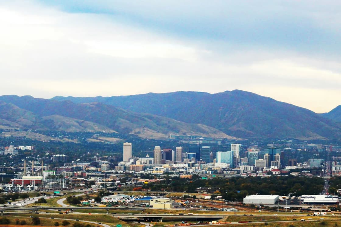 Downtown Salt Lake City's towers seen from a distance across the valley floor, with the Wasatch Range rising behind under a soft sky.
