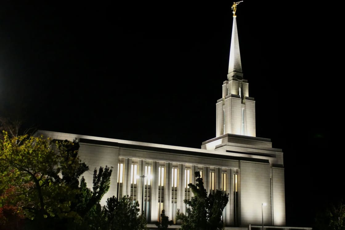 The Oquirrh Mountain Utah Temple in South Jordan lit up at night, its single spire and gold statue glowing above dark trees.