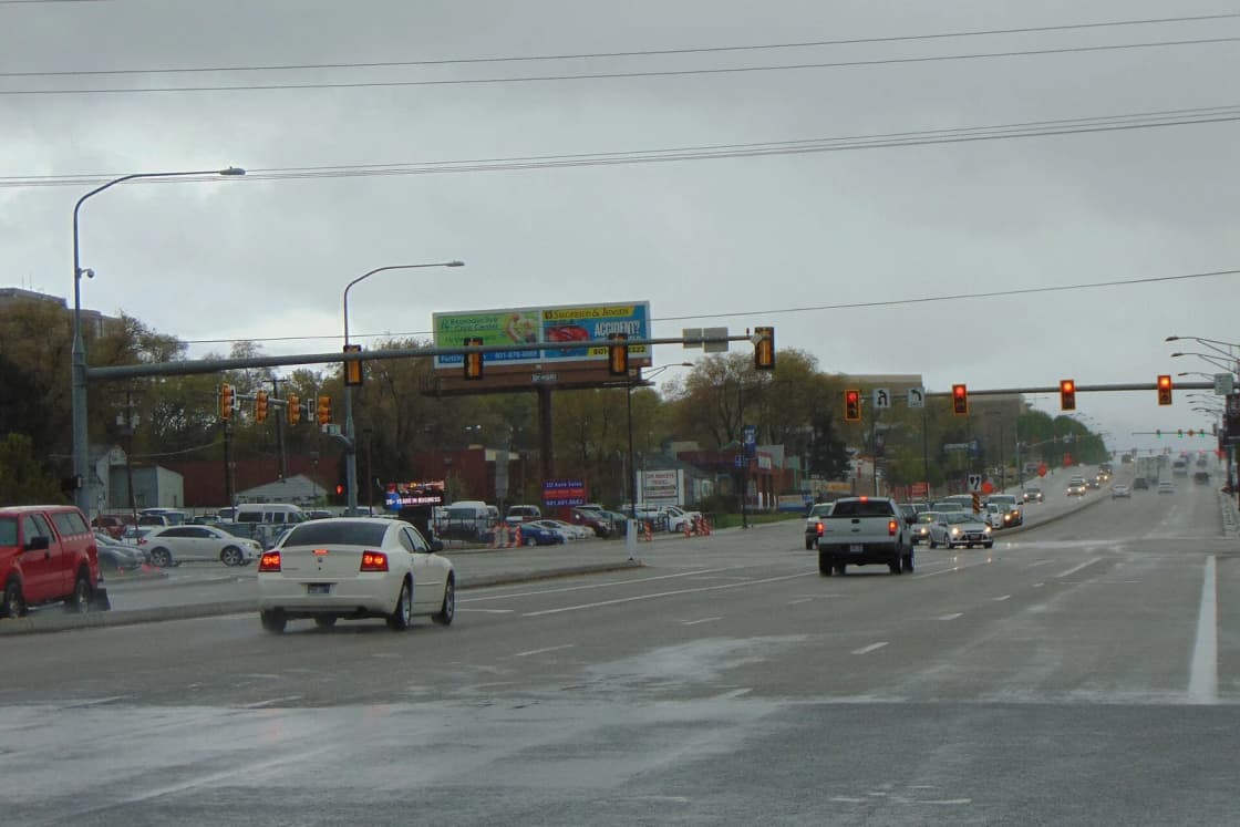 Cars head south on a wet State Street in Sandy on a gray afternoon, with traffic lights strung over the road.