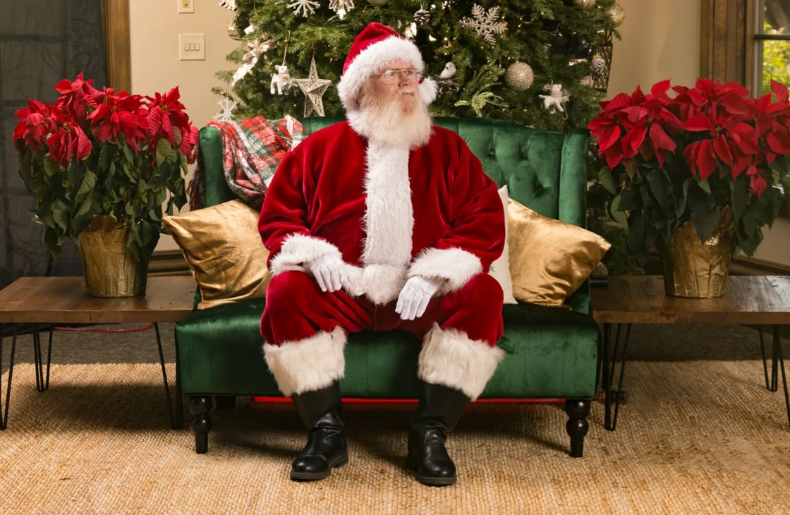 A white-bearded Santa in a red velvet suit sits on a green sofa flanked by red poinsettias with a decorated tree behind — Santa visits and events in Utah Valley