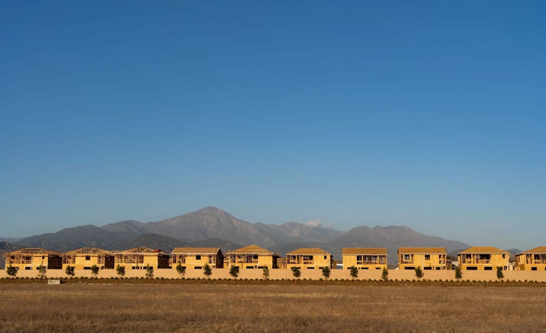 A row of new two-story houses stands on flat dry ground below a bare mountain under a clear sky