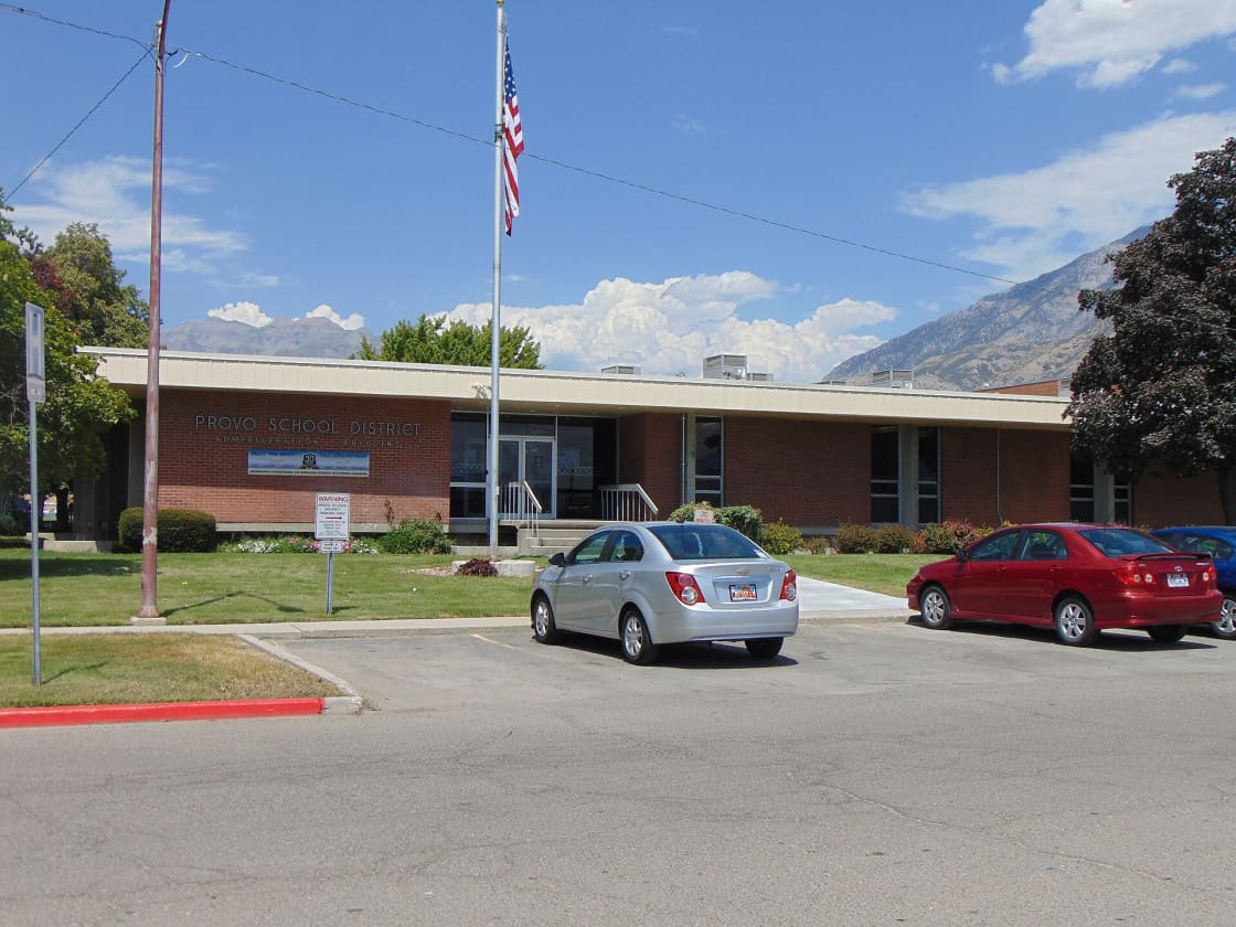 Single-story brick administration building lettered PROVO SCHOOL DISTRICT beneath a flagpole, with the Wasatch range rising behind it