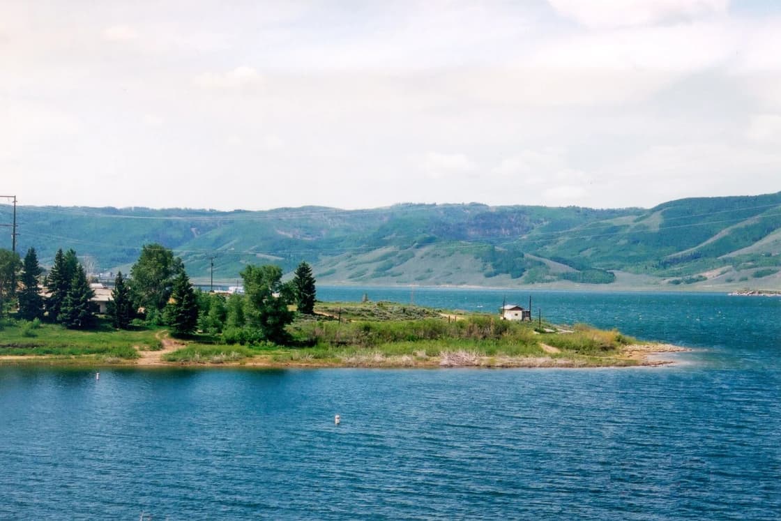 Blue water of Scofield Reservoir wraps a grassy point with pines, below green rolling hills.
