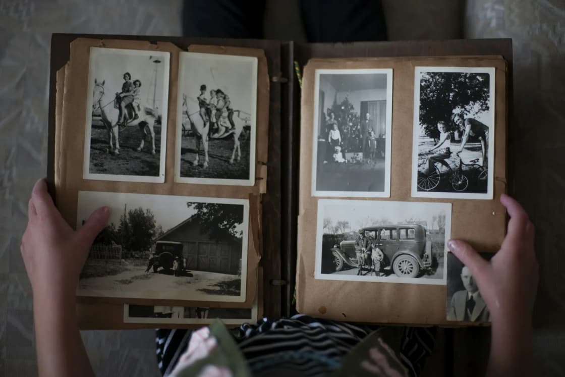 Hands hold open an old photo album of black-and-white family snapshots, including children on a pony and a vintage car.