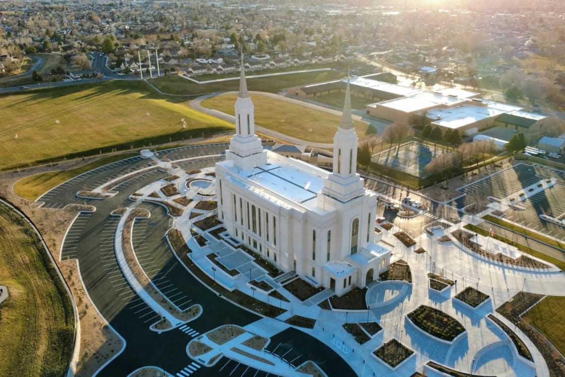 An aerial view of the white, twin-spired Lindon Utah Temple at golden hour, with its curving walkways and lawns and neighborhoods spreading beyond.