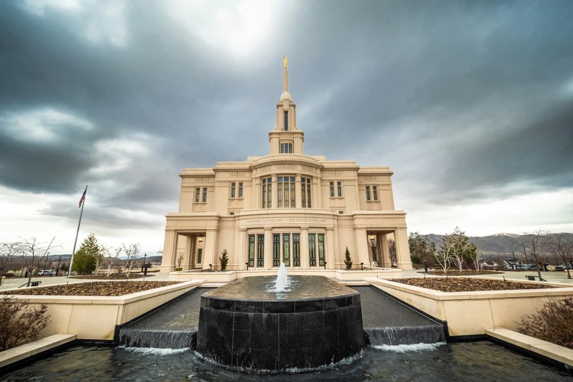 The cream-colored Payson Utah Temple under a dramatic cloudy sky, seen across the dark stone fountain and reflecting pool in front of it.