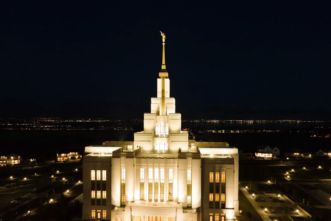 The Saratoga Springs Utah Temple glows white at night, with Utah Lake and distant city lights behind it.