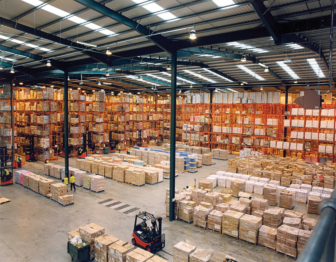 A warehouse worker in a high-visibility vest moving stacked cardboard boxes on a pallet trolley