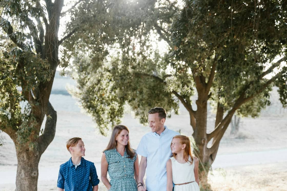 A family of four, two parents and a teenage son and daughter, walk together down a sunny dirt path under spreading oak trees.