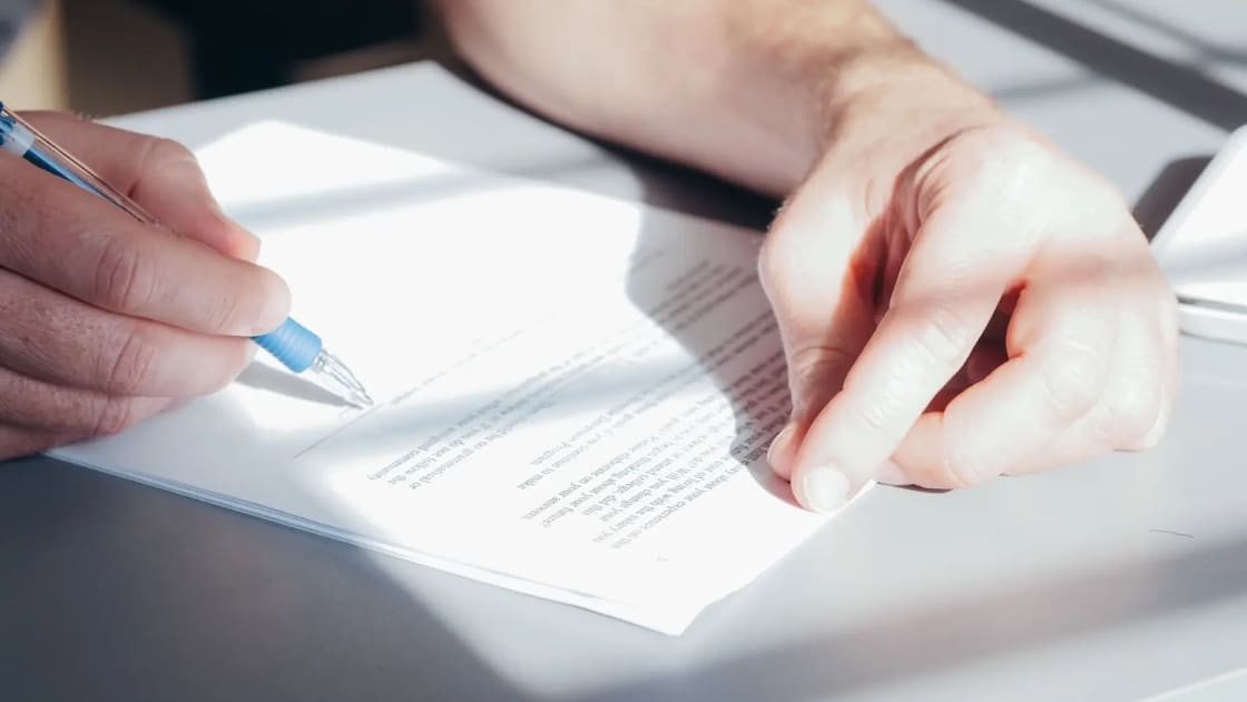 Two hands signing a printed document with a blue ballpoint pen on a bright desk