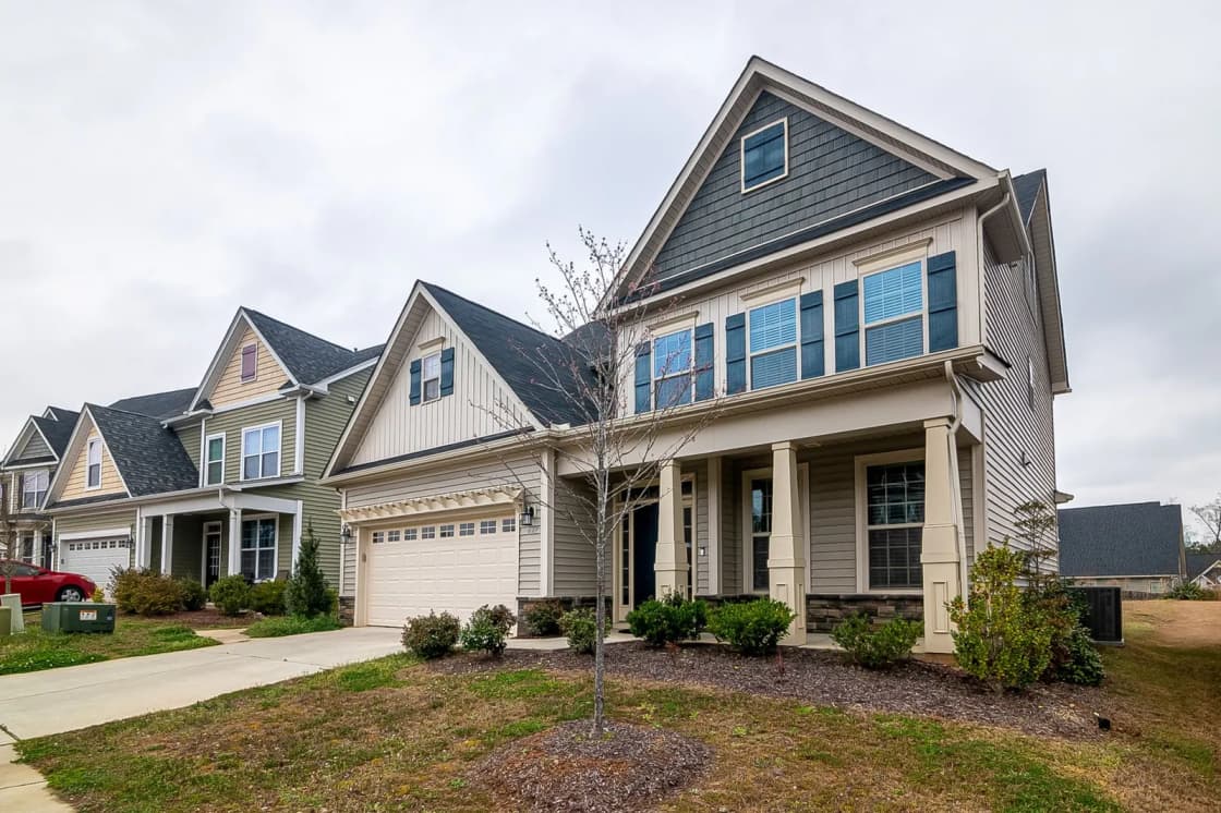 A row of two-story suburban houses with front porches, attached garages and young trees, under an overcast sky