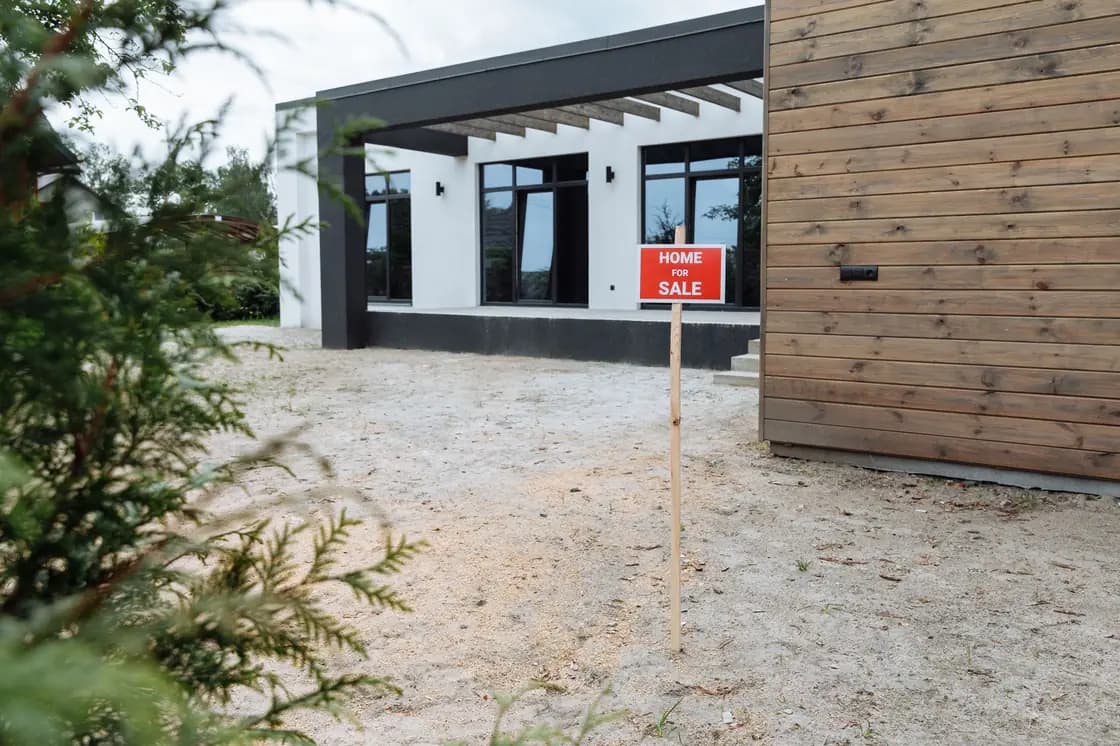 A red home-for-sale sign on a wooden stake standing in a bare sand forecourt in front of a house with dark-framed windows, a slatted pergola and a horizontal wood-clad wall