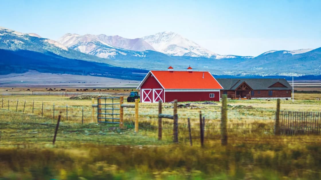 A red barn and a log house on open pasture behind a wooden fence and a metal gate, with snow-dusted mountains in the distance