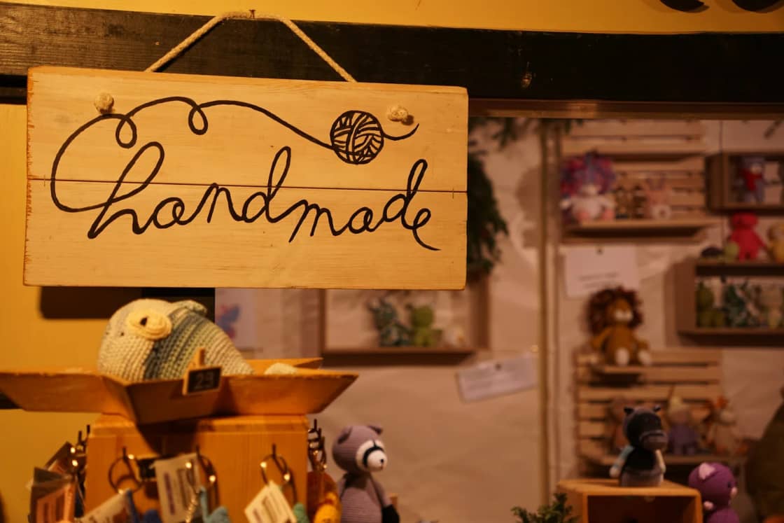 A hand-lettered wooden sign reading handmade hangs over a maker's booth stocked with crocheted animals and small gifts.