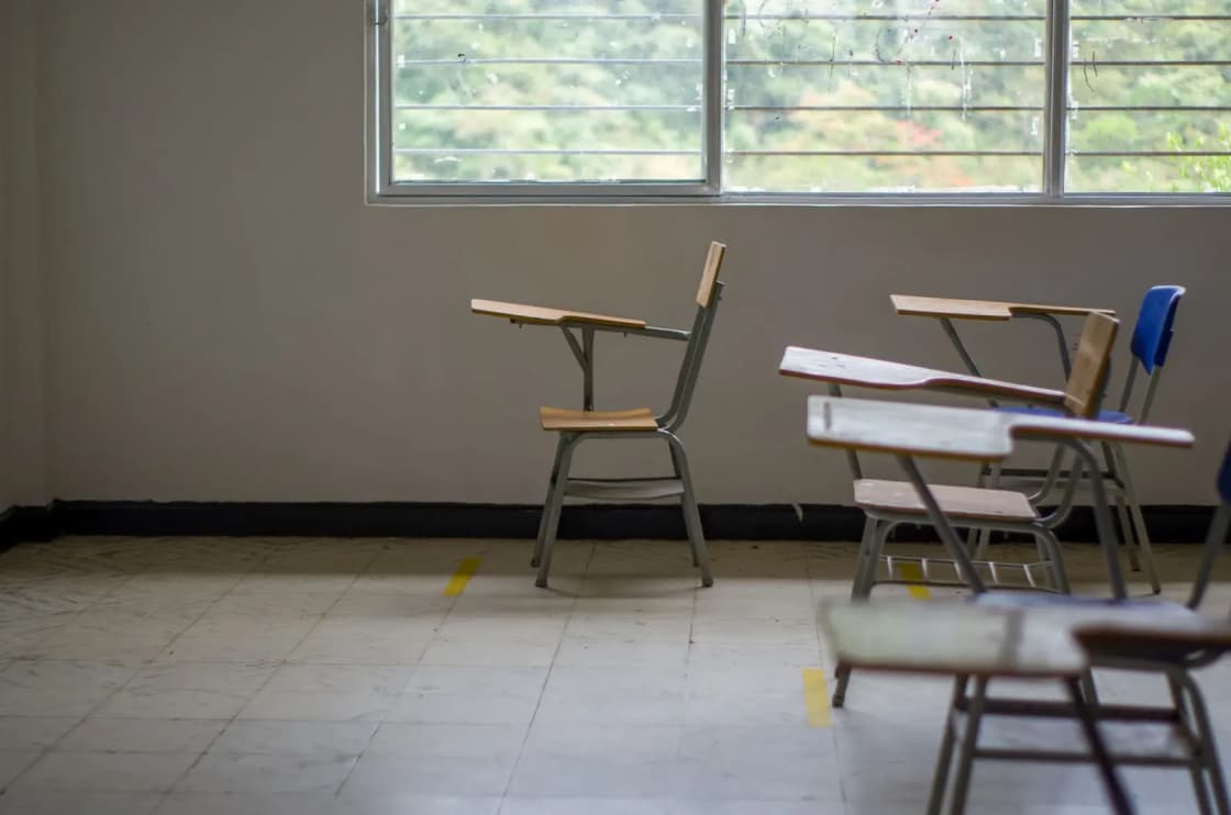 Empty tablet-arm desk chairs scattered across a tiled classroom floor beneath a long window