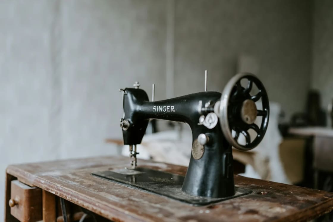 A vintage black Singer sewing machine with gold lettering sits on a worn wooden treadle table in a dim workroom.