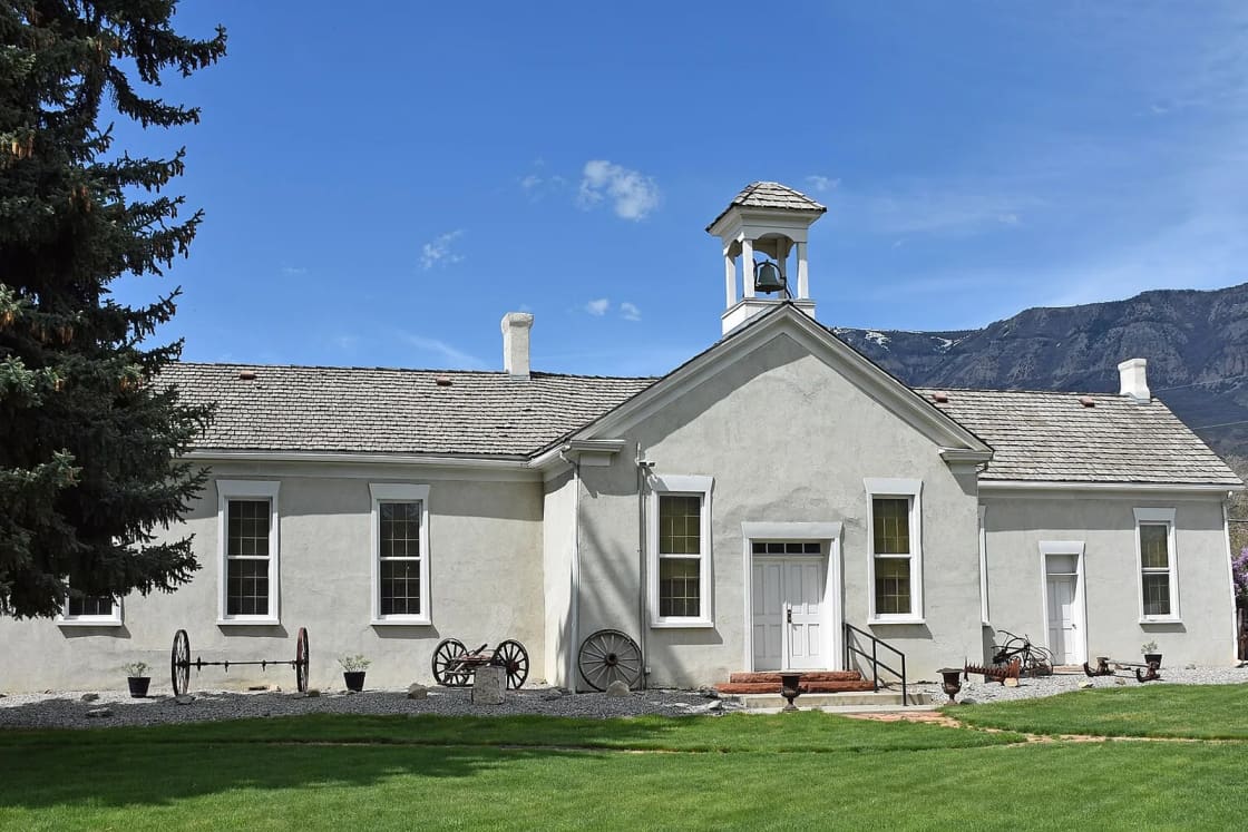 Pleasant Grove's white Old Bell School with its bell tower and green door, wagon wheels lining the lawn, a tall evergreen at left and the mountains rising behind