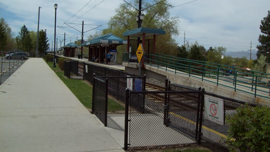 The platform at Historic Sandy Station, where UTA's Route 994 ski bus to Snowbird and Alta begins, with teal-roofed TRAX shelters, overhead wires and a fenced walkway on a green spring day