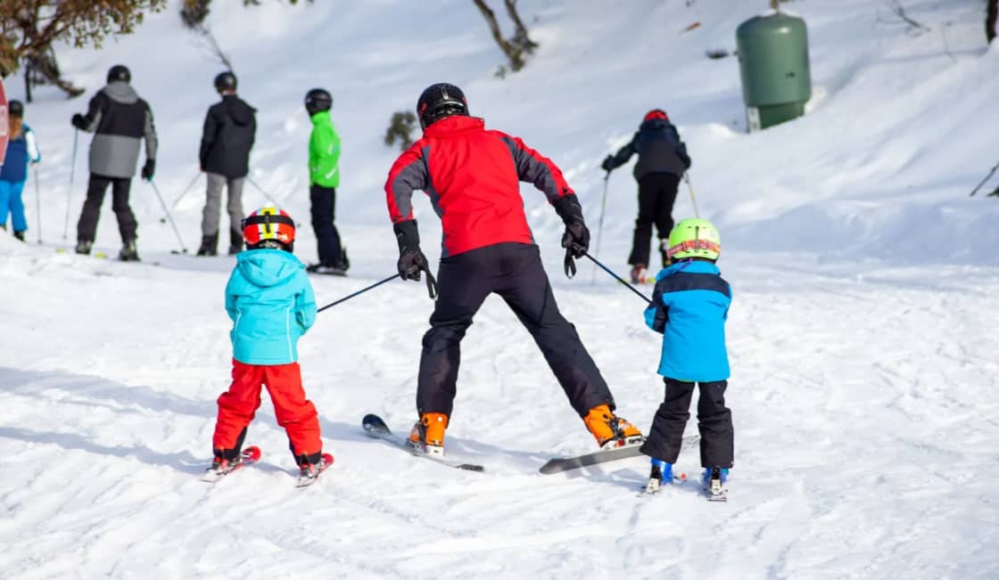 A parent in a red jacket skis between two small children in blue, all three snowplowing down a gentle beginner slope with other skiers behind — choosing a family ski pass in Utah Valley
