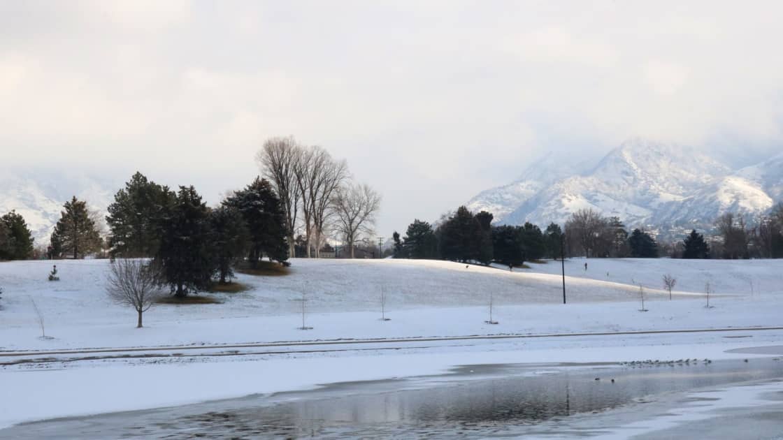 Sugar House Park in Salt Lake City under fresh snow, its rolling white hills dotted with evergreens and bare trees above a partly frozen pond with waterfowl, and snowy Wasatch peaks in the clouds behind