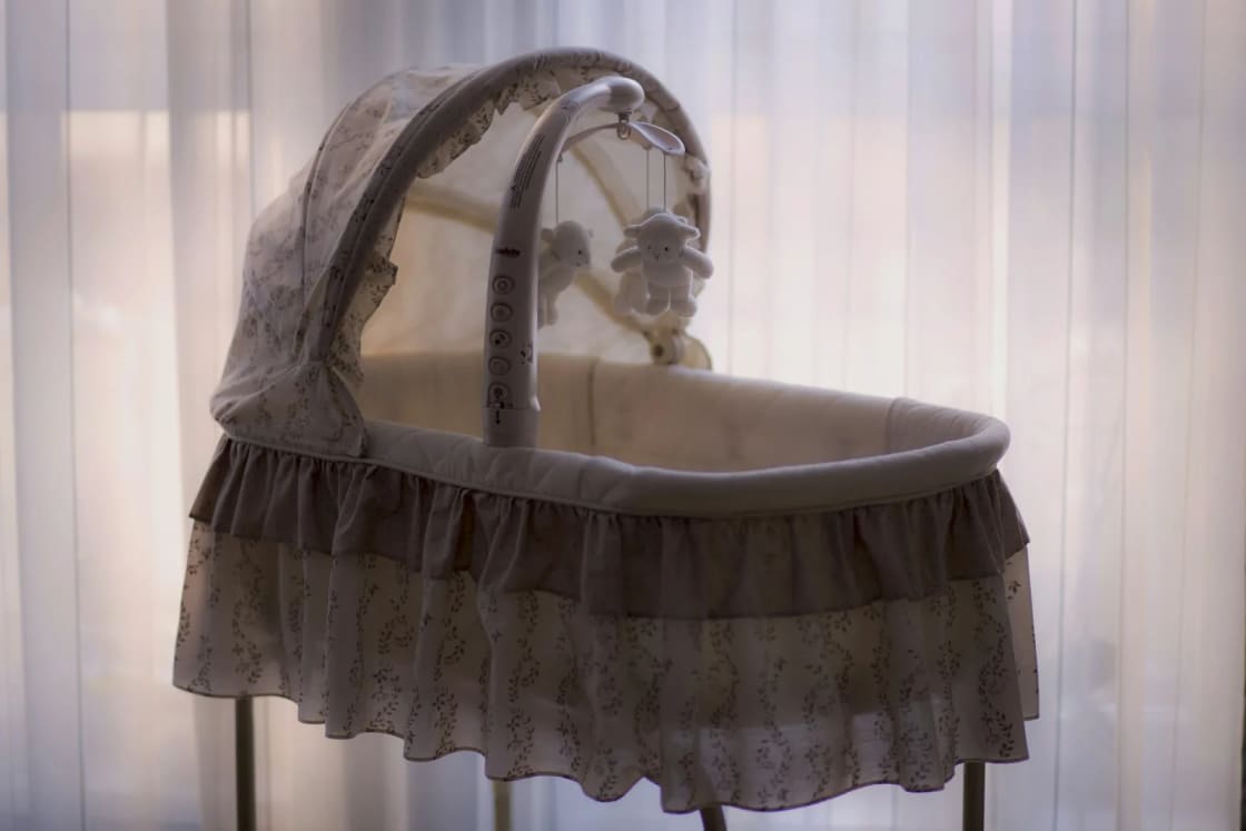An empty bassinet with a ruffled skirt and a small hanging mobile stands in front of a curtained window in soft light.