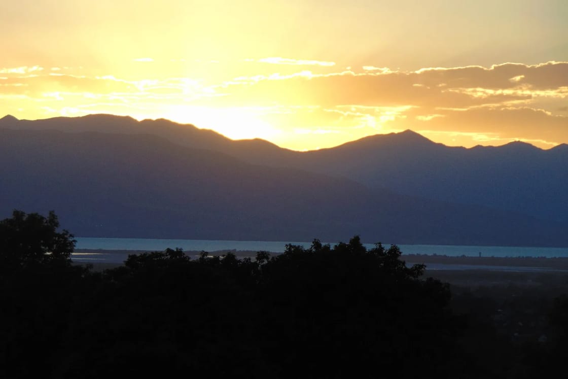 The sun sets behind the Lake Mountains across Utah Lake, with dark trees in the foreground.