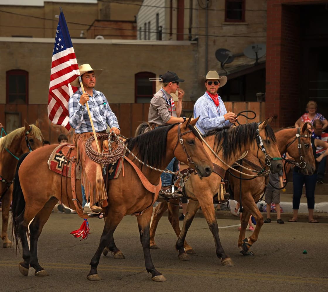 Riders on horseback carrying an American flag along a small-town parade route as spectators watch from the curb