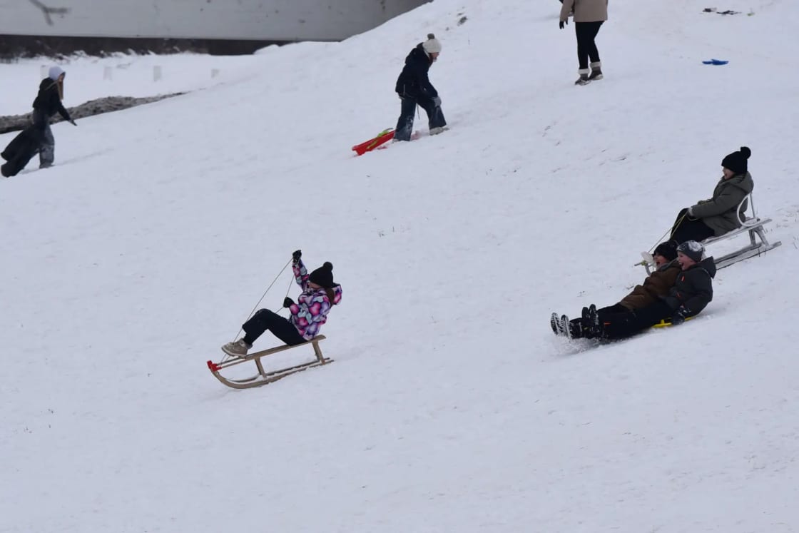 Sledders sliding down a broad snowy hill on a gray winter day while another person hauls a sled back up the slope
