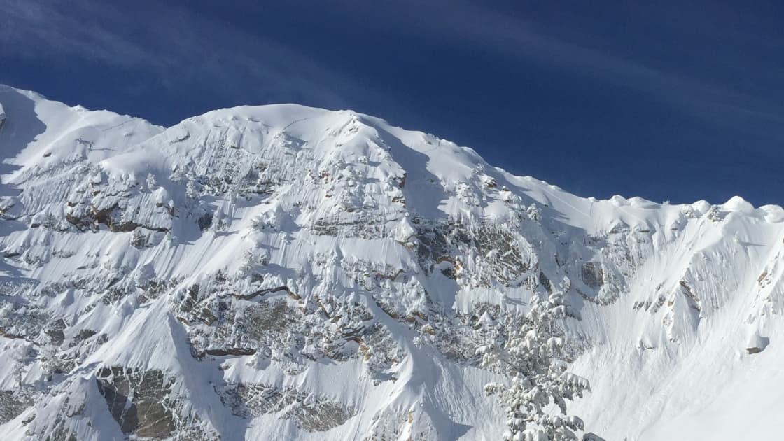 A rugged, snow-plastered peak above Snowbasin under a deep blue winter sky streaked with thin cloud, with the edge of a snow-covered evergreen in the lower corner