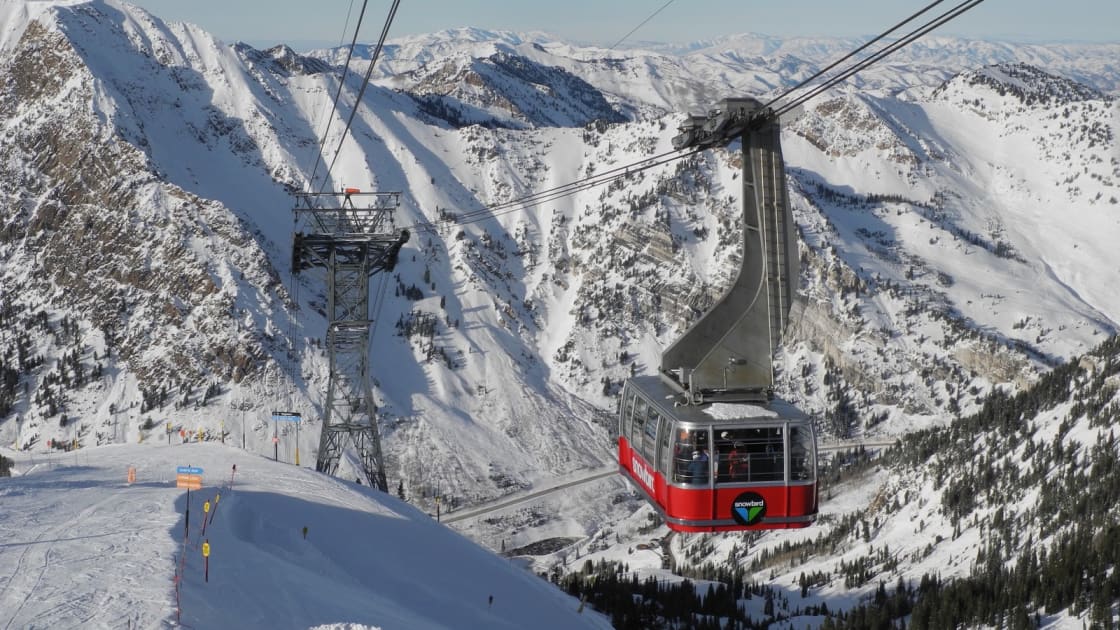 Snowbird's red Aerial Tram car swinging out from the summit station on Hidden Peak, cables stretching over snow-covered ridges, chutes and forested slopes under a clear sky