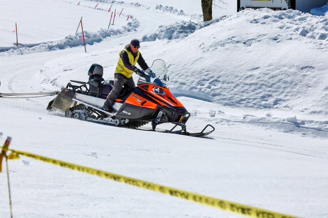 A rider in a yellow jacket and helmet drives an orange snowmobile across packed snow, with a strip of yellow tape in the foreground and snowy hills behind