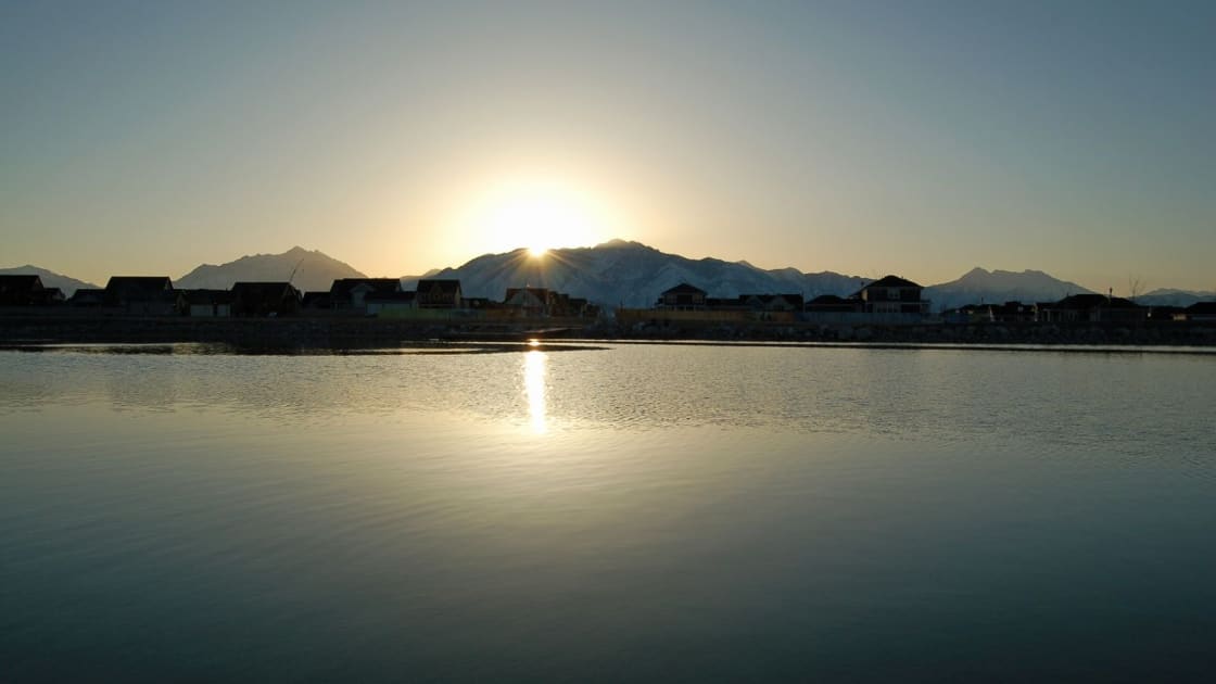 The sun rising over the Wasatch Mountains behind the far shore of Oquirrh Lake in Daybreak, with rooftops in silhouette and a line of light reflected on the still water
