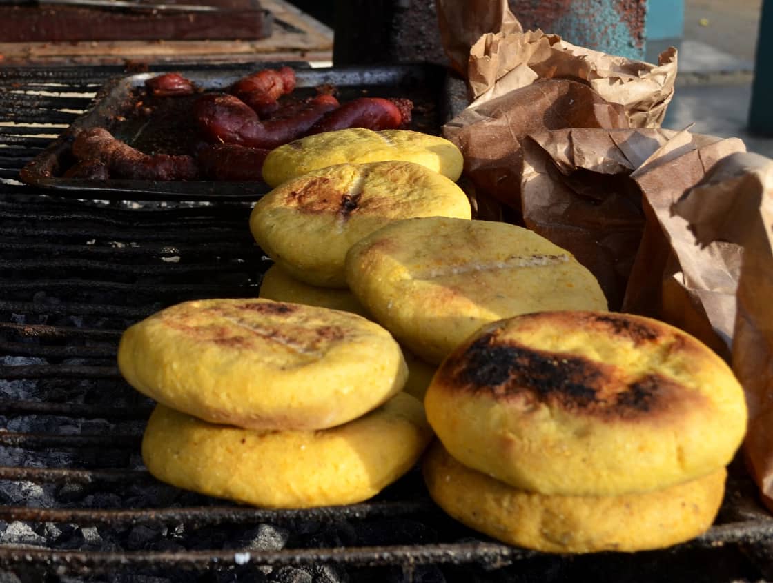 Thick yellow arepas browning on a charcoal grill beside a tray of sausages