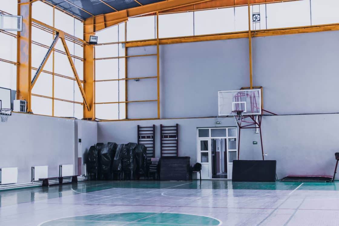 An empty indoor sports hall with a green and gray painted court, a wall-mounted basketball hoop and yellow steel roof trusses overhead