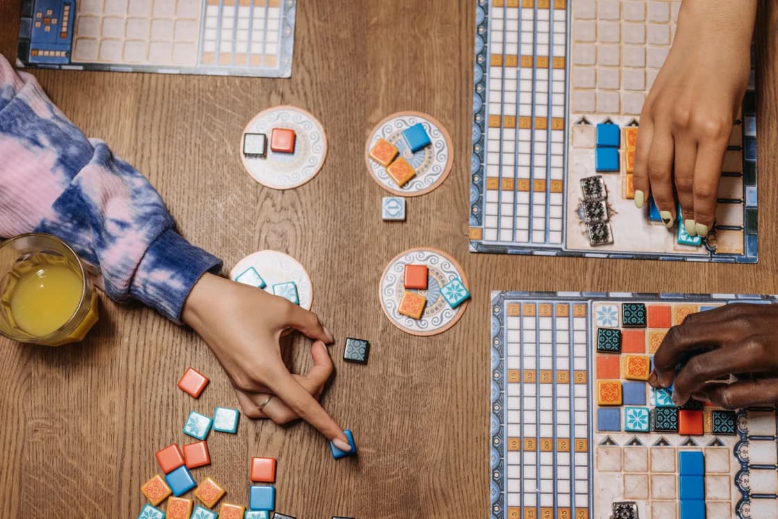 Hands of several players moving tiles on a colorful board game on a wooden table.