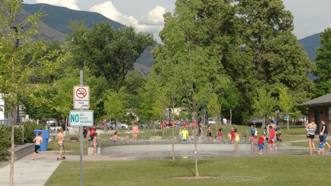 The splash pad in Springville on a summer day, children playing among the fountains in the distance beside a lawn, shade trees and a park building, with mountains behind