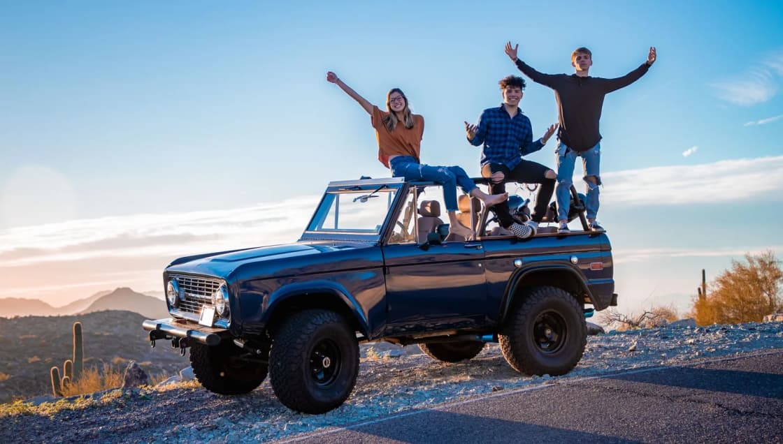Four friends stand on the roof and hood of a blue off-road truck on a desert road at dusk, arms raised, with low hills and a pale sky behind them