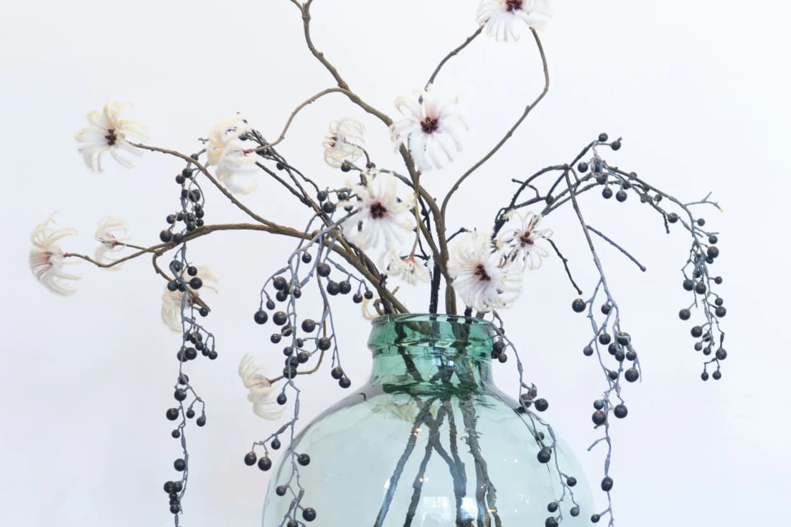 Bare branches with small white flowers and dark berries arranged in a round green glass jug on a stool against a white wall.