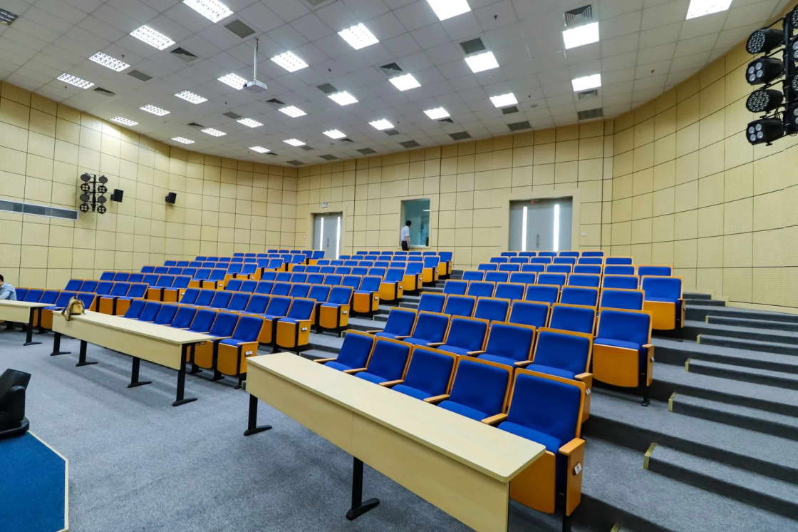 An empty tiered lecture hall with rows of blue seats and wooden desks, lit but with no students seated