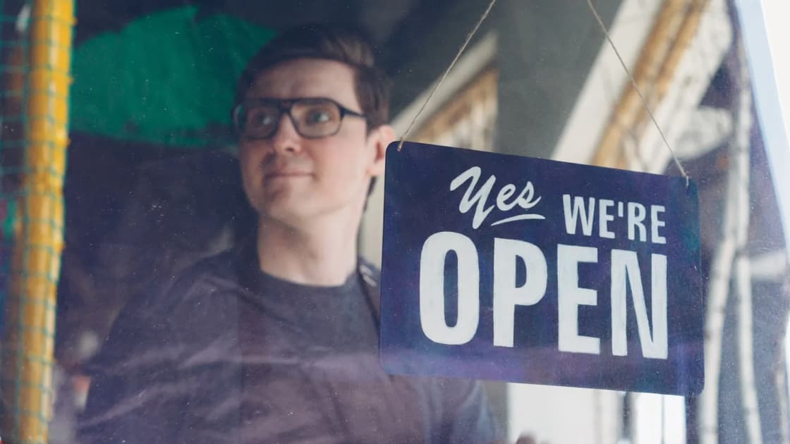 A young shop owner in an apron seen through a glass door, standing beside a hanging sign that reads Yes we're open