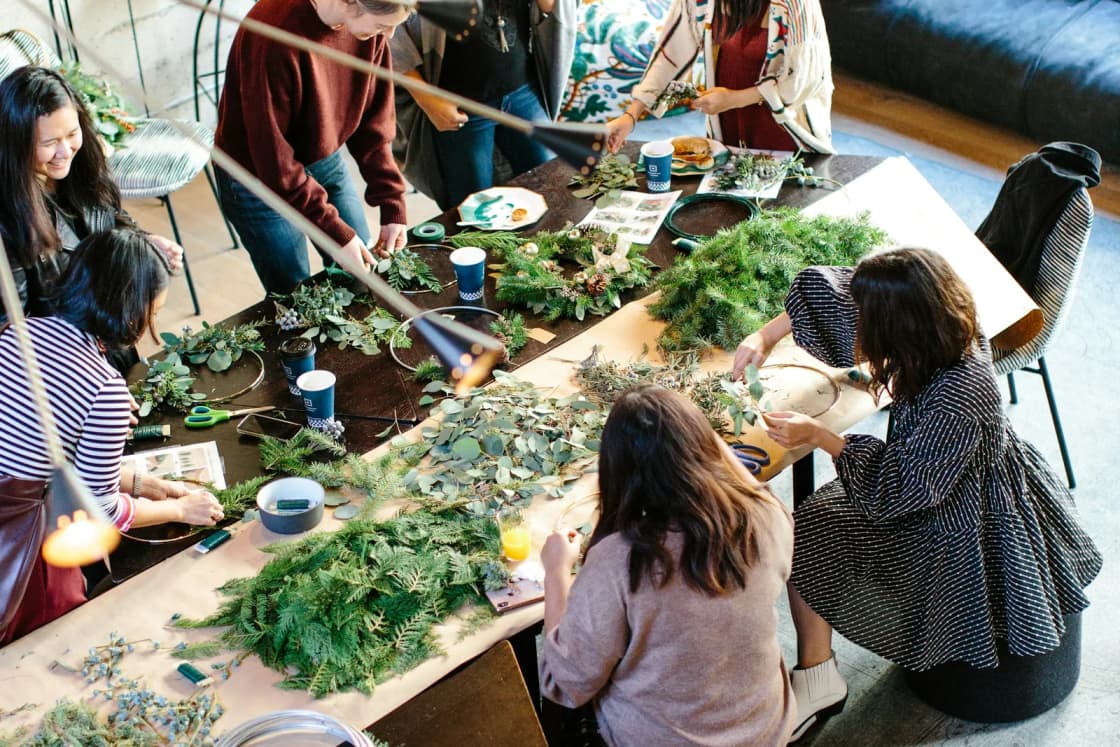 A group of friends gathers around a long table covered in fresh greenery, making wreaths together in a bright loft.