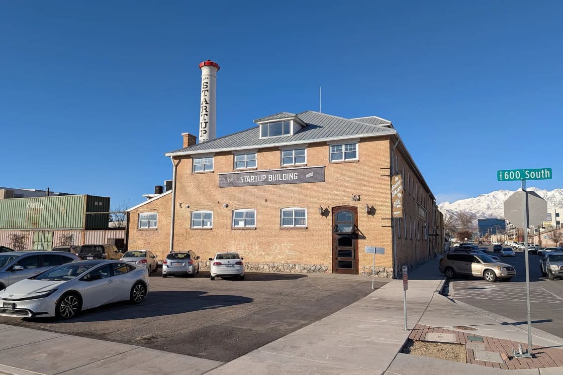 The two-story brick Startup Building in Provo with its tall painted smokestack, a parking lot and snowy peaks behind.