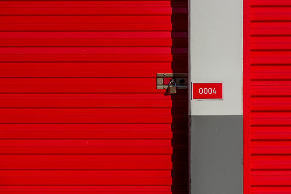 A bright red roll-up storage unit door secured with a padlock, the unit number 0004 on a small sign beside it