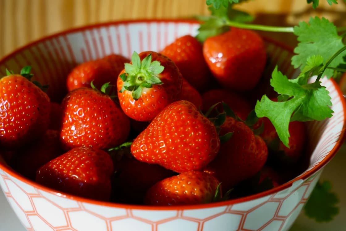 A bowl of ripe red strawberries with a patterned orange and white rim, with green herb leaves resting on top