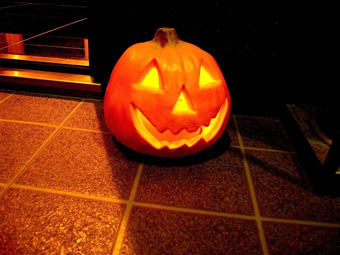 A carved jack-o'-lantern lit from inside with a candle, sitting on a tiled doorstep at night, its triangular eyes and jagged grin glowing orange against the dark