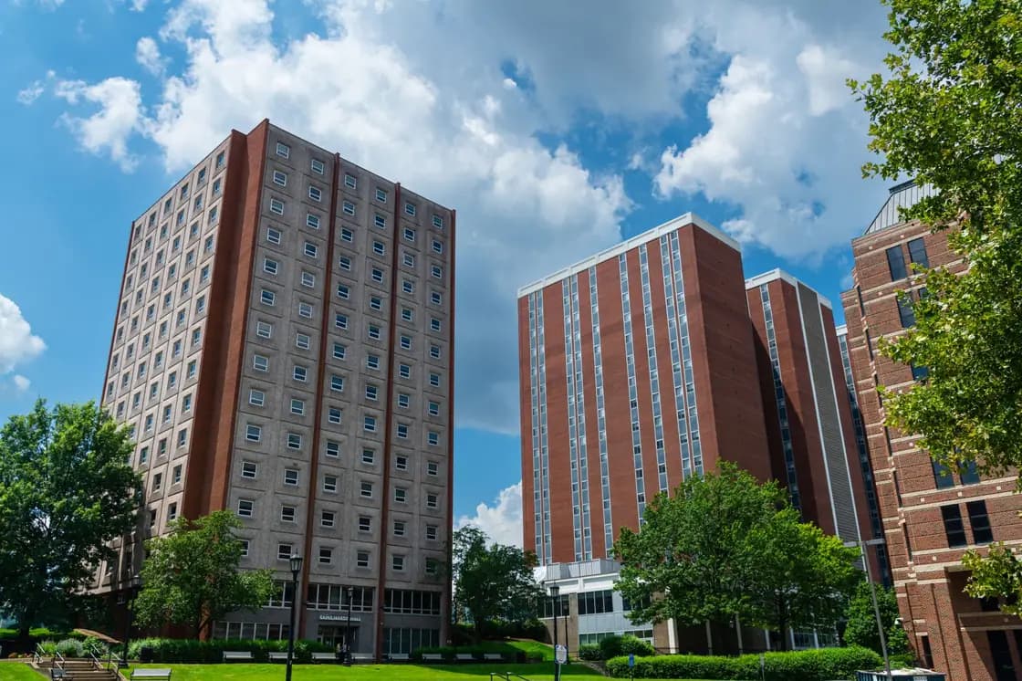 Two tall residential tower blocks, one pale concrete with a grid of small windows and one red brick, seen across a lawn with mature trees under a partly clouded sky