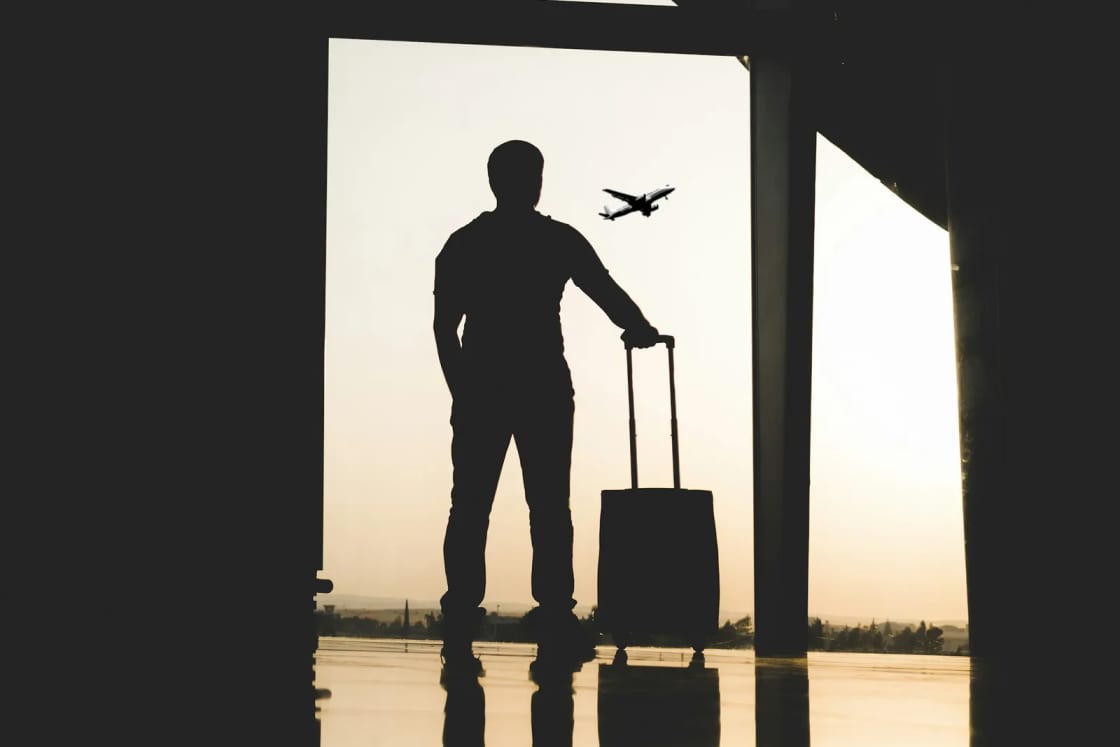 The silhouette of a traveler standing with a rolling suitcase at a tall airport window, a jet climbing into a pale evening sky outside