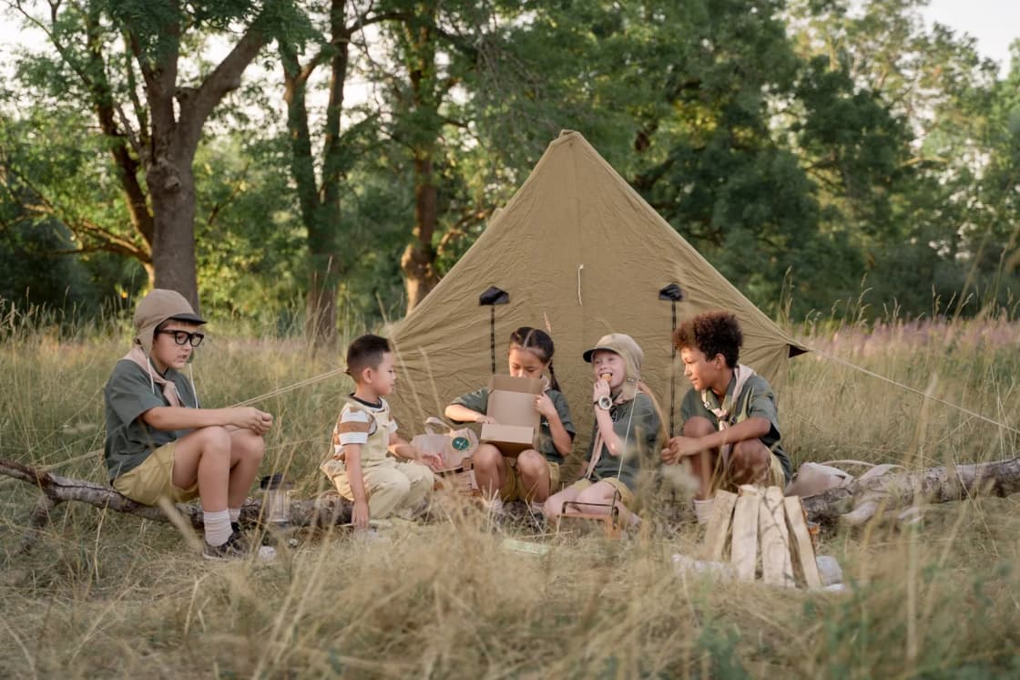 Five children in green camp shirts and neckerchiefs sit on a log in tall golden grass in front of a canvas tent, one wearing a paper hat and another holding a camp stove kettle