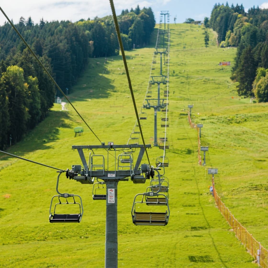 Empty chairlift chairs hang from a cable above a bright green grassy ski run in summer, with dark conifer forest along both sides and a second lift line climbing the slope behind